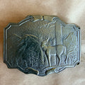 A rectangular metal belt buckle with an embossed deer in a forest landscape, shown on a light brown textured background.