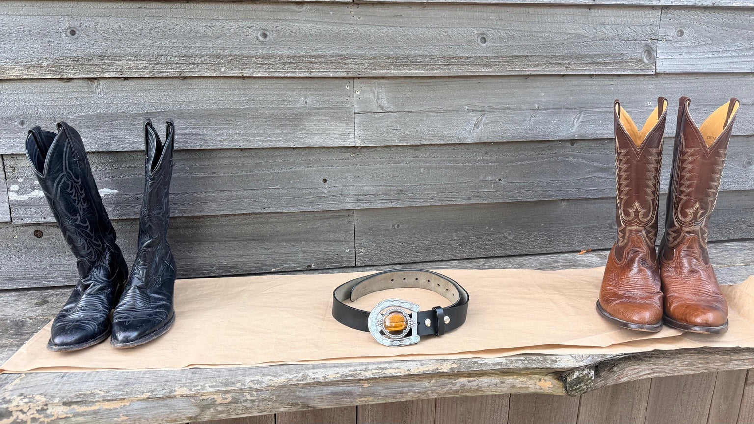 Two pairs of western boots and a western belt arranged on a tan paper sheet against a gray wooden plank background. The left pair is black, the center is a black belt with a silver buckle, and the right pair is brown with tan interiors.